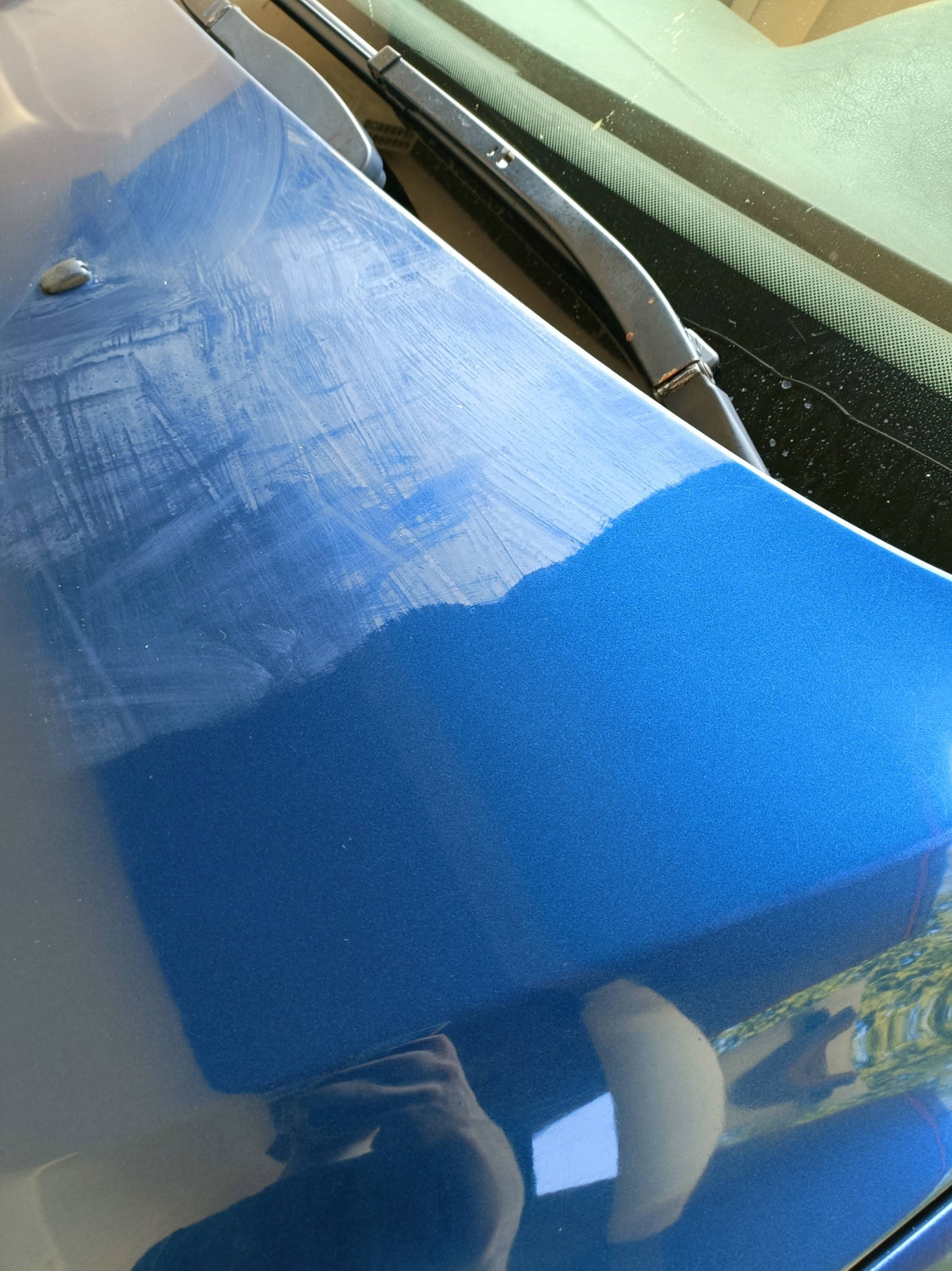 Fragment niebieskiej karoserii auta z widocznymi śladami polerowania, ukazujący różnicę w połysku przed i po zabiegu. W tle wycieraczka szyby.