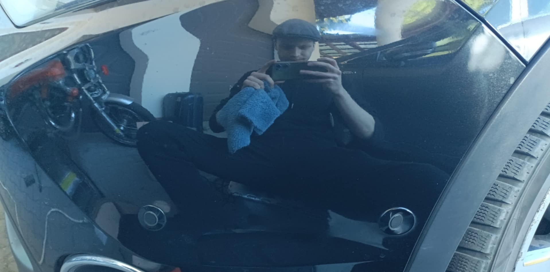 Błyszcząca karoseria czarnego samochodu odbija postać mężczyzny z telefonem i niebieską ściereczką, obok widoczny motocykl. Detailing auta.