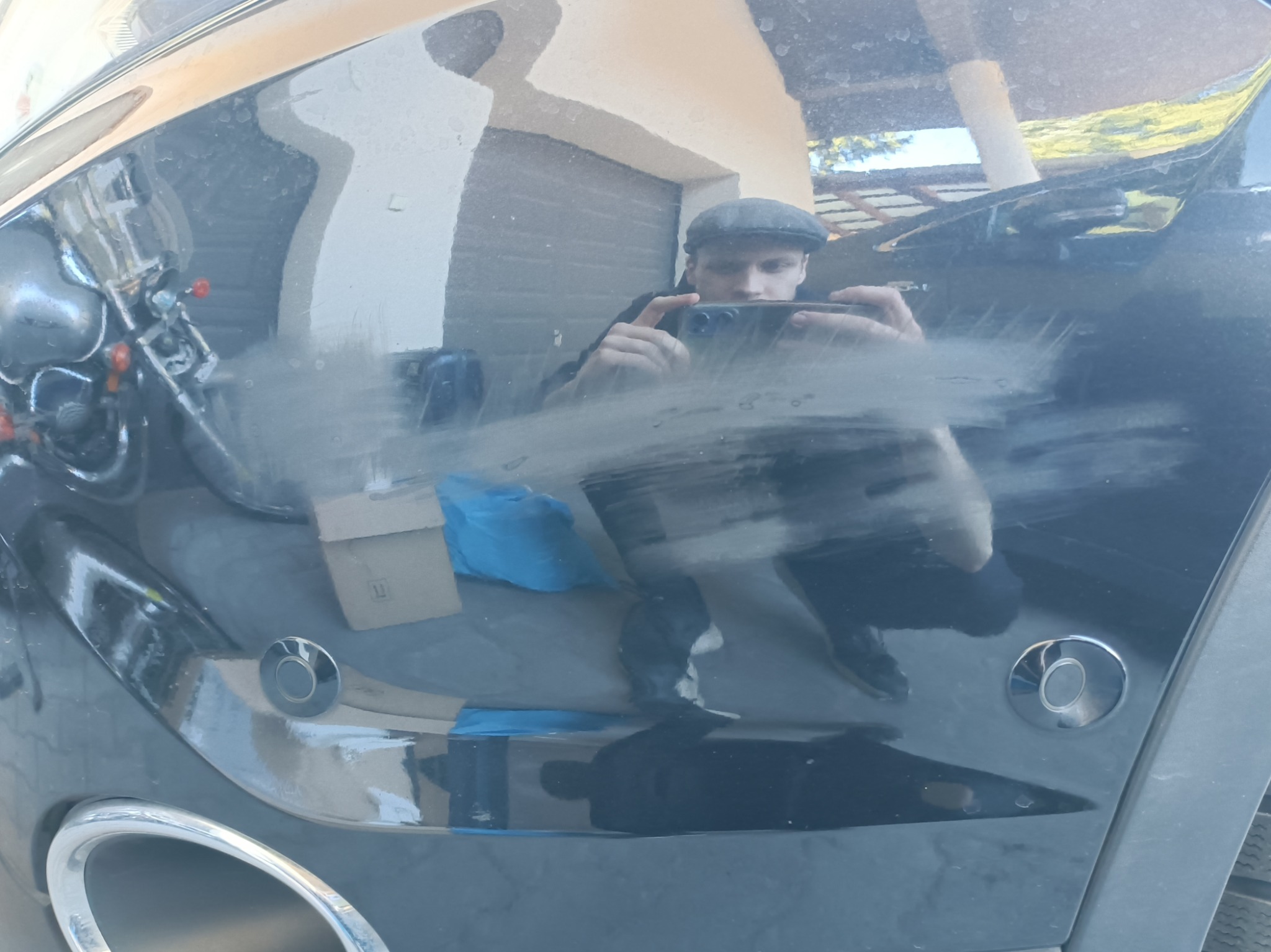 Brudny, czarny samochód z widocznymi smugami i odbiciem osoby robiącej zdjęcie telefonem. Widoczne czujniki parkowania i fragment opony.