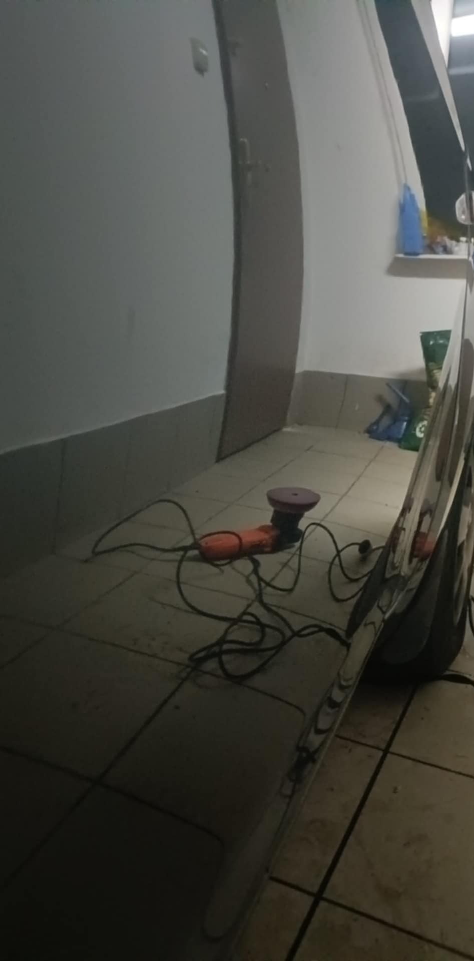 Polerka samochodowa leży na podłodze obok błyszczącego, wypolerowanego elementu karoserii w pomieszczeniu. Widoczne środki czystości na półce w tle.