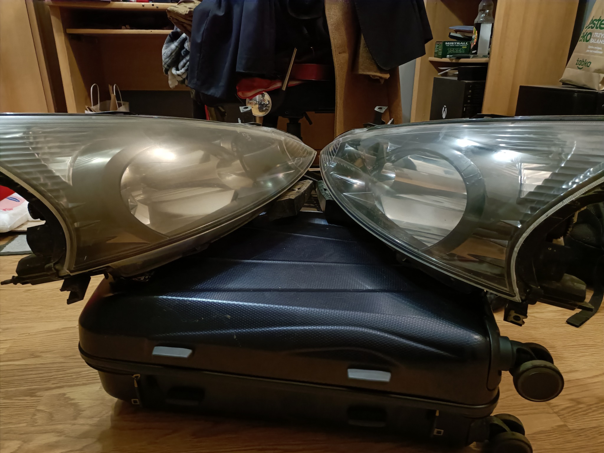 Dwie lampy samochodowe, jedna matowa, druga czysta, leżące na czarnej walizce na kółkach. Widoczne tło z półkami i przedmiotami.