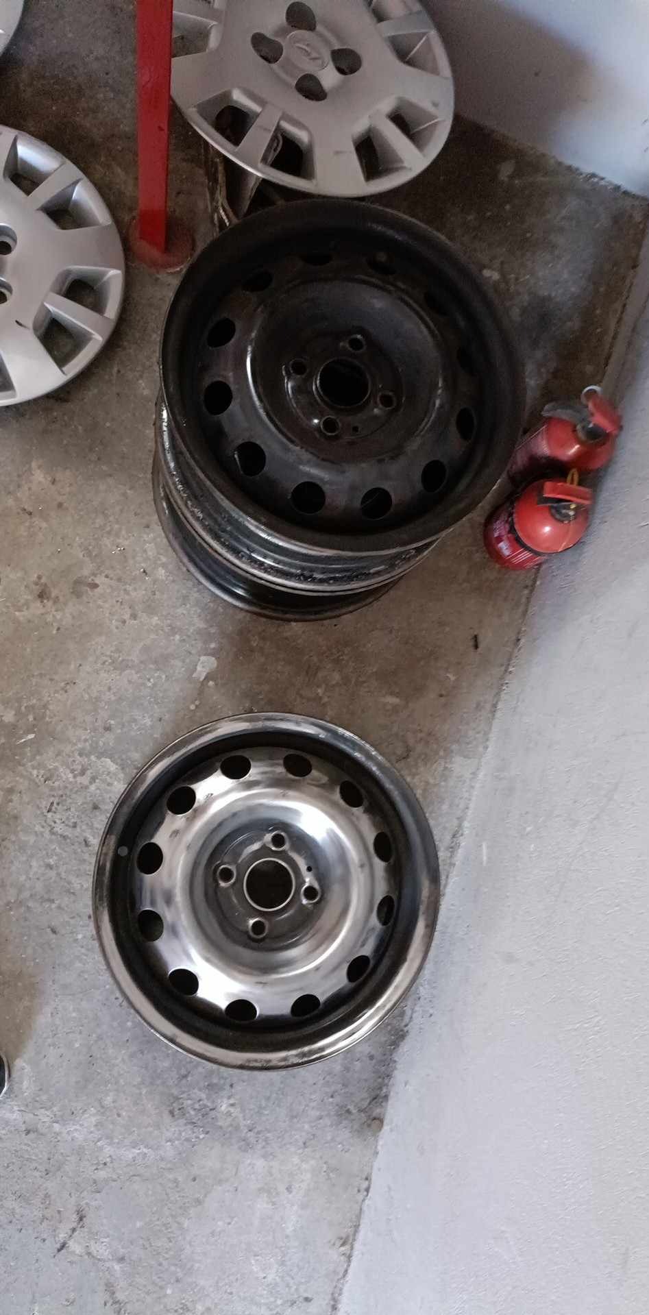 Stos felg stalowych, jedna czysta, inne czarne, obok kołpaki i gaśnice. Felgi po czyszczeniu prezentują się lepiej.