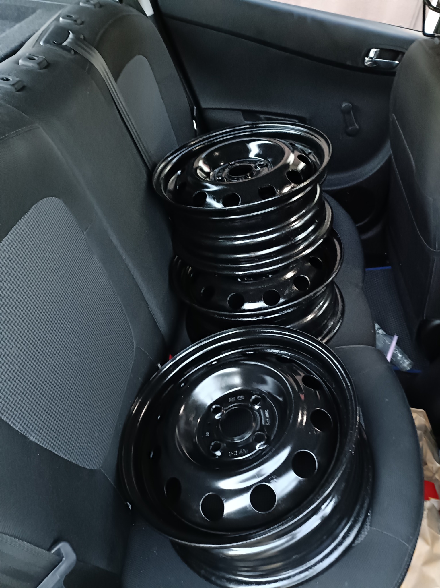 Stos czarnych, stalowych felg samochodowych ułożonych na tylnym siedzeniu pojazdu. Widoczne detale tapicerki i uchwyt drzwi.