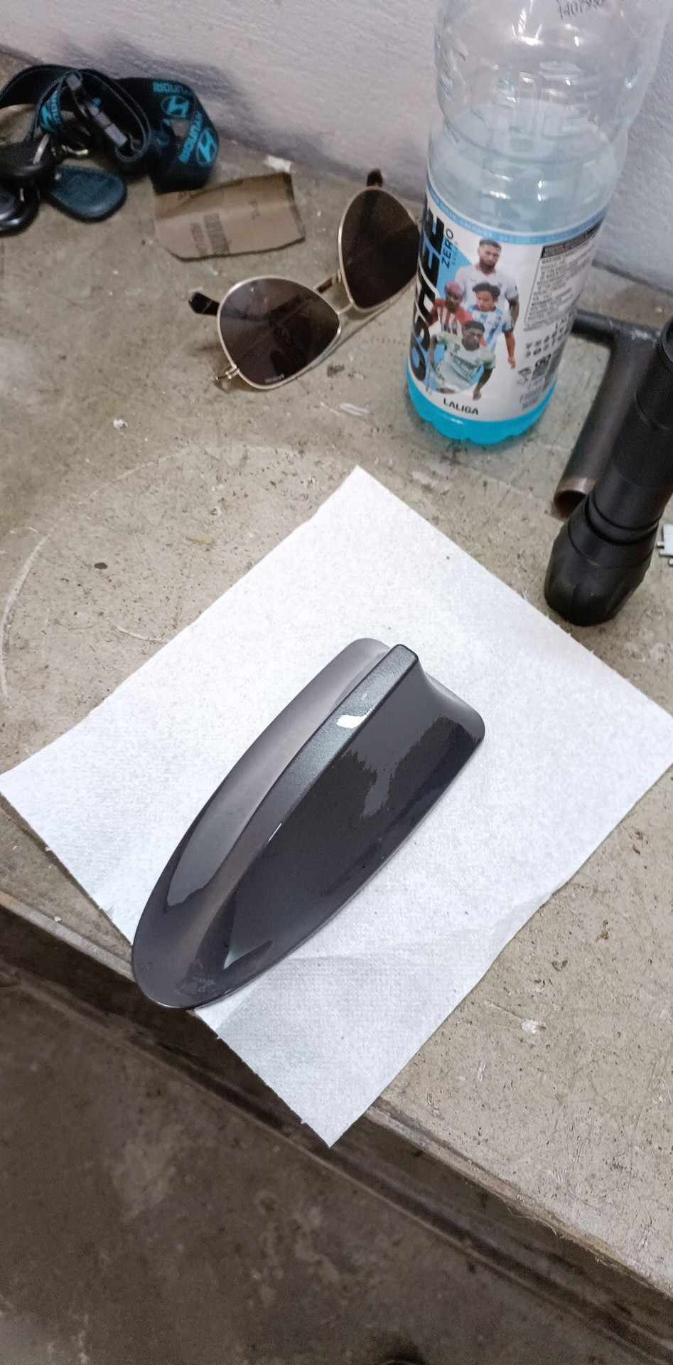 Błyszcząca, szara antena samochodowa typu płetwa rekina leży na białej chusteczce na tle warsztatowego stołu z okularami i butelką wody.