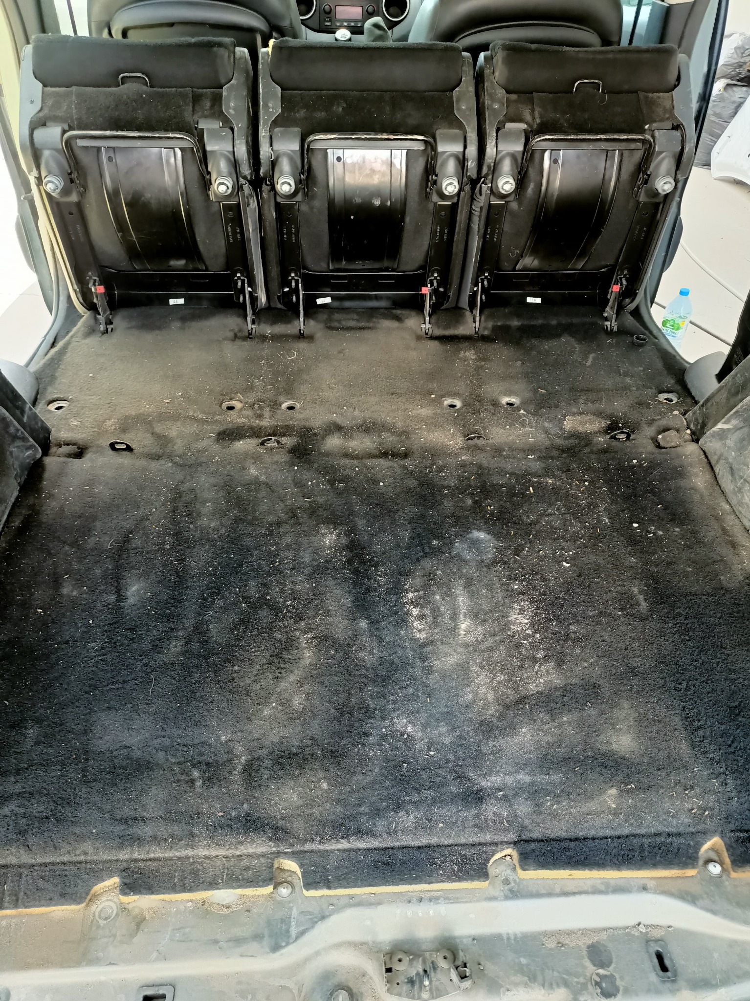 Wnętrze samochodu z wyjętymi siedzeniami, ukazujące brudną tapicerkę podłogową przed czyszczeniem. Widoczne mocowania siedzeń i butelka wody w tle.