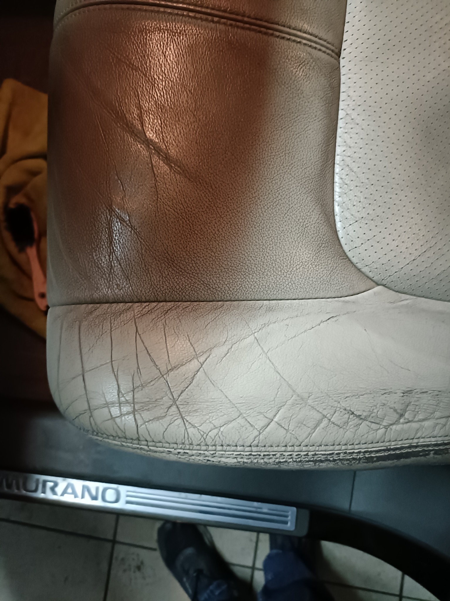Fragment skórzanego fotela samochodowego w odcieniach beżu i brązu, widoczne ślady użytkowania na dolnej części, napis 'MURANO' na progu.