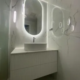 Nowoczesna łazienka z owalnym lustrem LED, białą umywalką nablatową i szafką z ryflowanym frontem na tle marmurowych płytek.