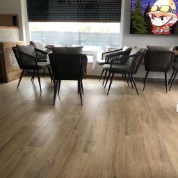 Juja - Jasne panele podłogowe w salonie z ciemnymi krzesłami i roletami w oknie. Na ścianie wisi telewizor z animowaną postacią. Widoczne fragmenty mebli i okna.