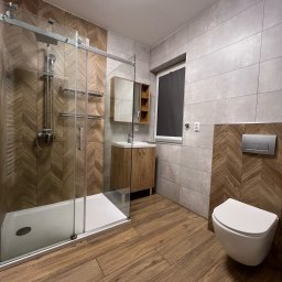 Juja - Nowoczesna łazienka po remoncie: kabina prysznicowa typu walk-in z drewnopodobnymi płytkami, wisząca toaleta, drewniana szafka z umywalką, szare płytki.