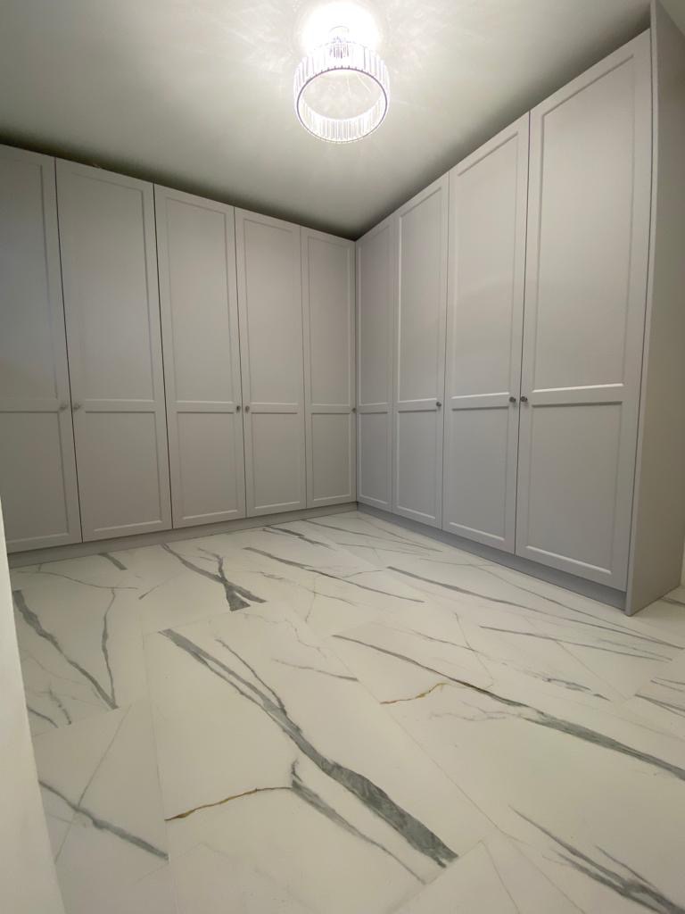 Jasne wnętrze garderoby z szarymi szafami w zabudowie i podłogą z białych płytek imitujących marmur. Centralnie umieszczony, okrągły plafon.