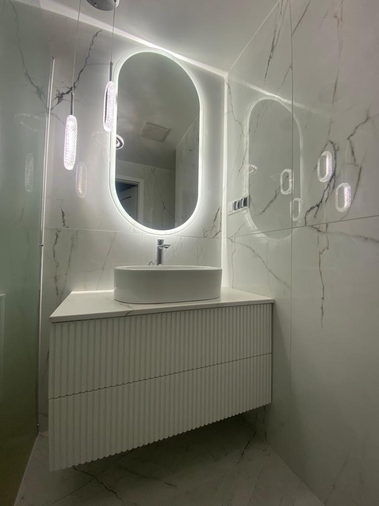 Nowoczesna łazienka z owalnym lustrem LED, białą umywalką nablatową i szafką z ryflowanym frontem na tle marmurowych płytek.