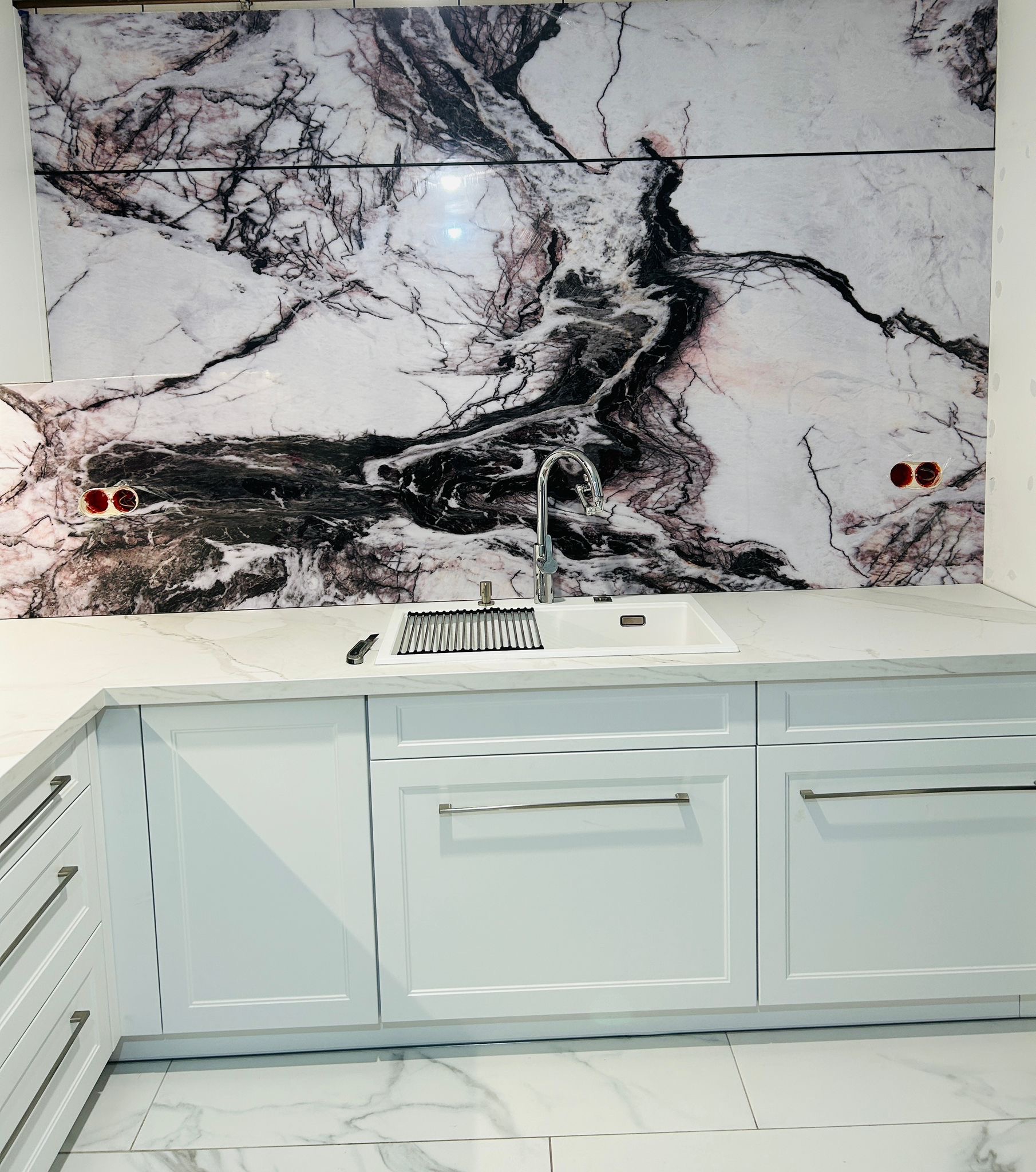Nowoczesna kuchnia z białymi szafkami, marmurowym blatem i oryginalnym panelem ściennym o wzorze marmuru z czarnymi żyłami. Umywalka ze stali nierdzewnej.