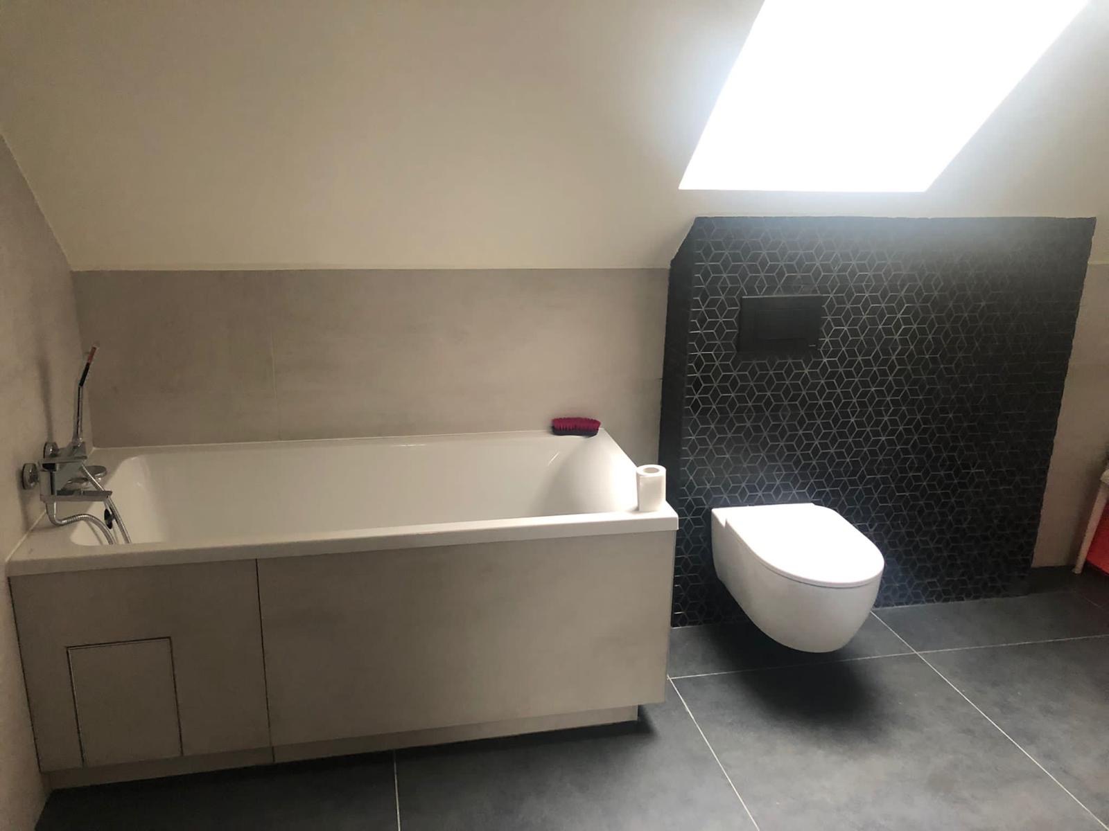 Wykończona łazienka na poddaszu ze skośnym sufitem, z zabudowaną wanną, wiszącą toaletą i czarną ścianą z geometrycznym wzorem. Widoczne szare płytki podłogowe.