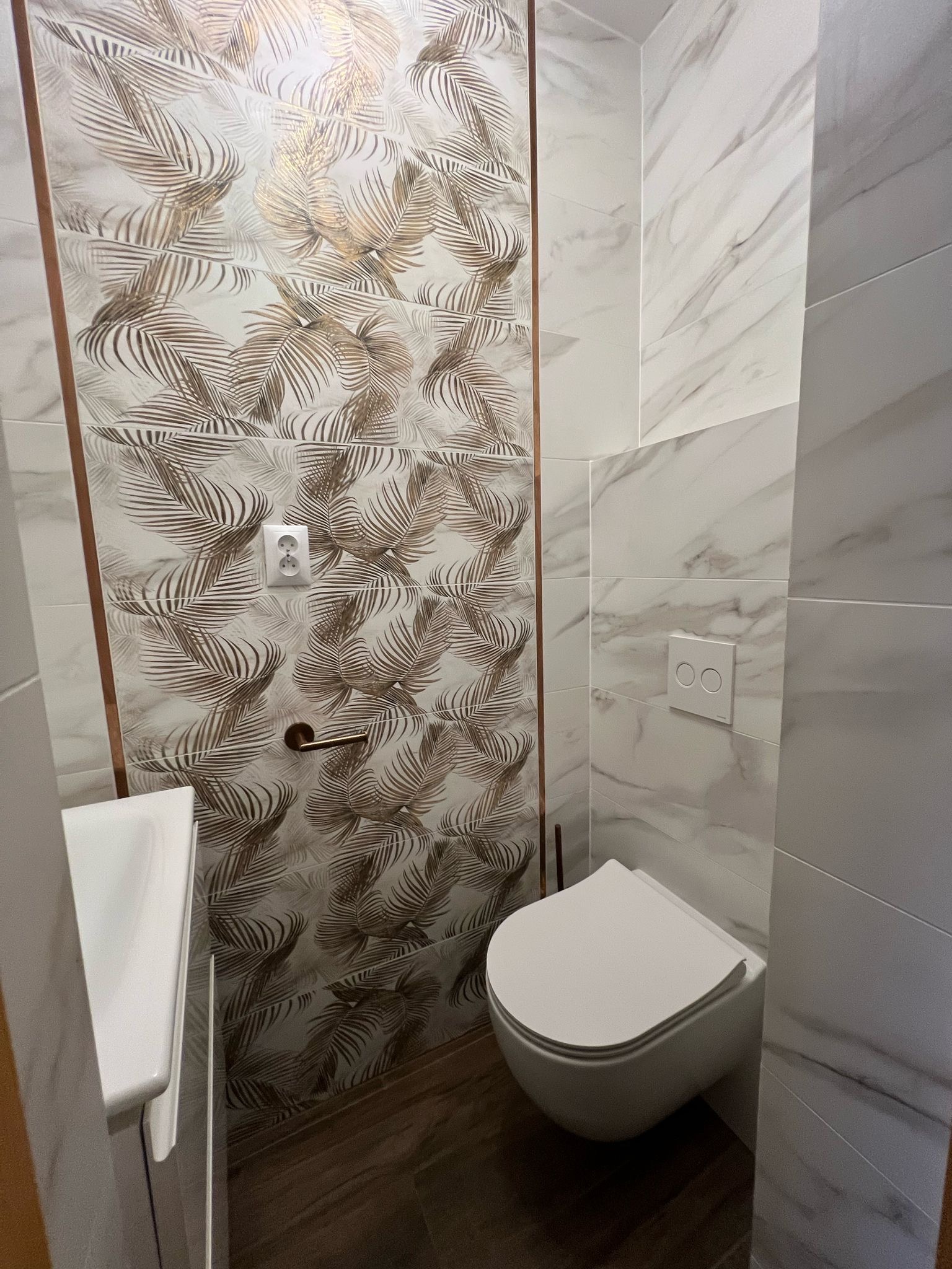 Wykończona łazienka z marmurowymi i wzorzystymi płytkami, wisząca toaleta, umywalka. Nowoczesny design z akcentami złota i brązu.