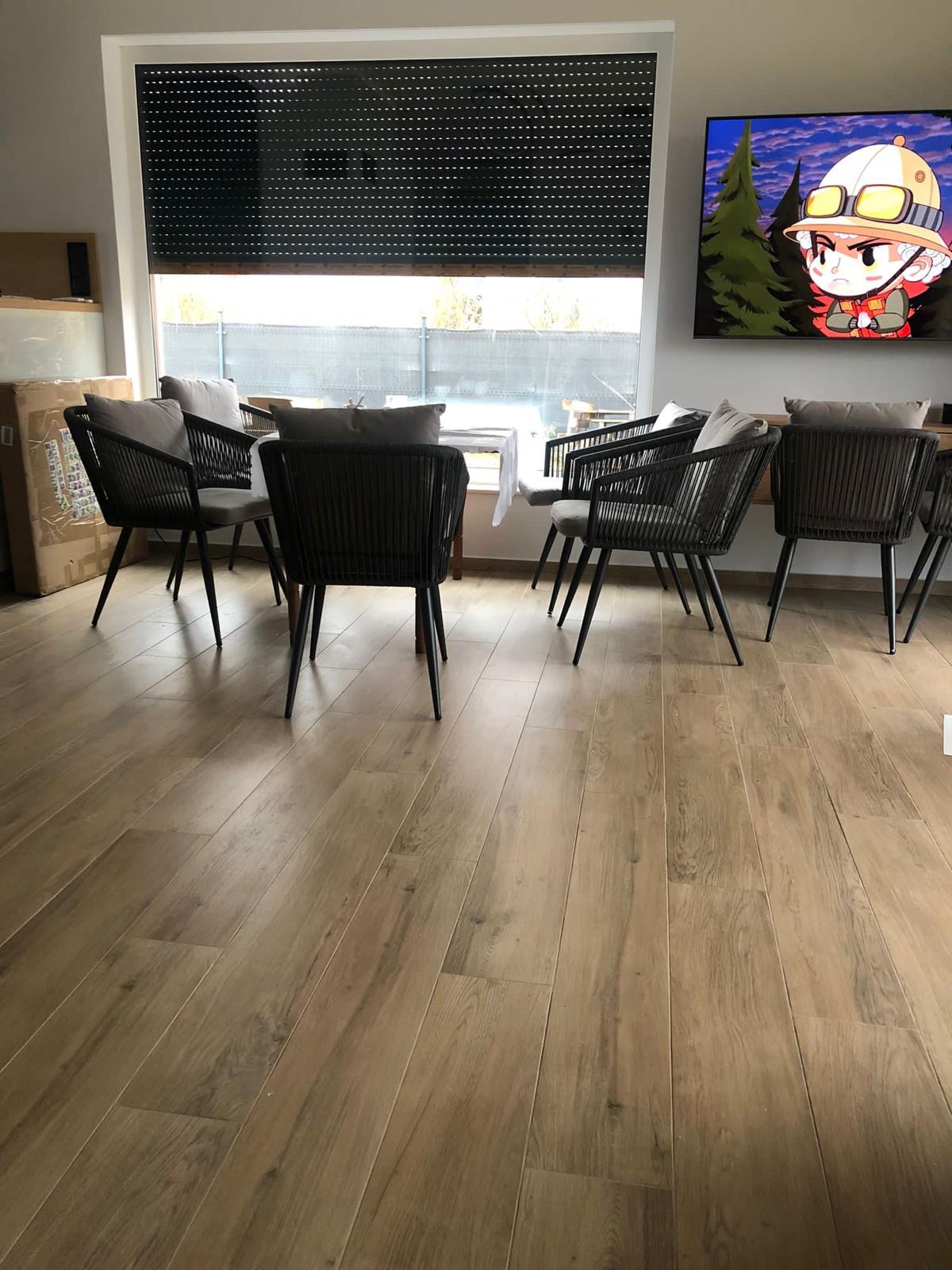Jasne panele podłogowe w salonie z ciemnymi krzesłami i roletami w oknie. Na ścianie wisi telewizor z animowaną postacią. Widoczne fragmenty mebli i okna.