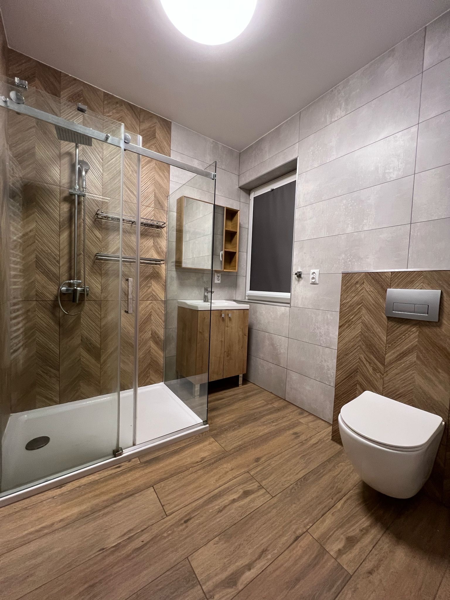 Nowoczesna łazienka po remoncie: kabina prysznicowa typu walk-in z drewnopodobnymi płytkami, wisząca toaleta, drewniana szafka z umywalką, szare płytki.