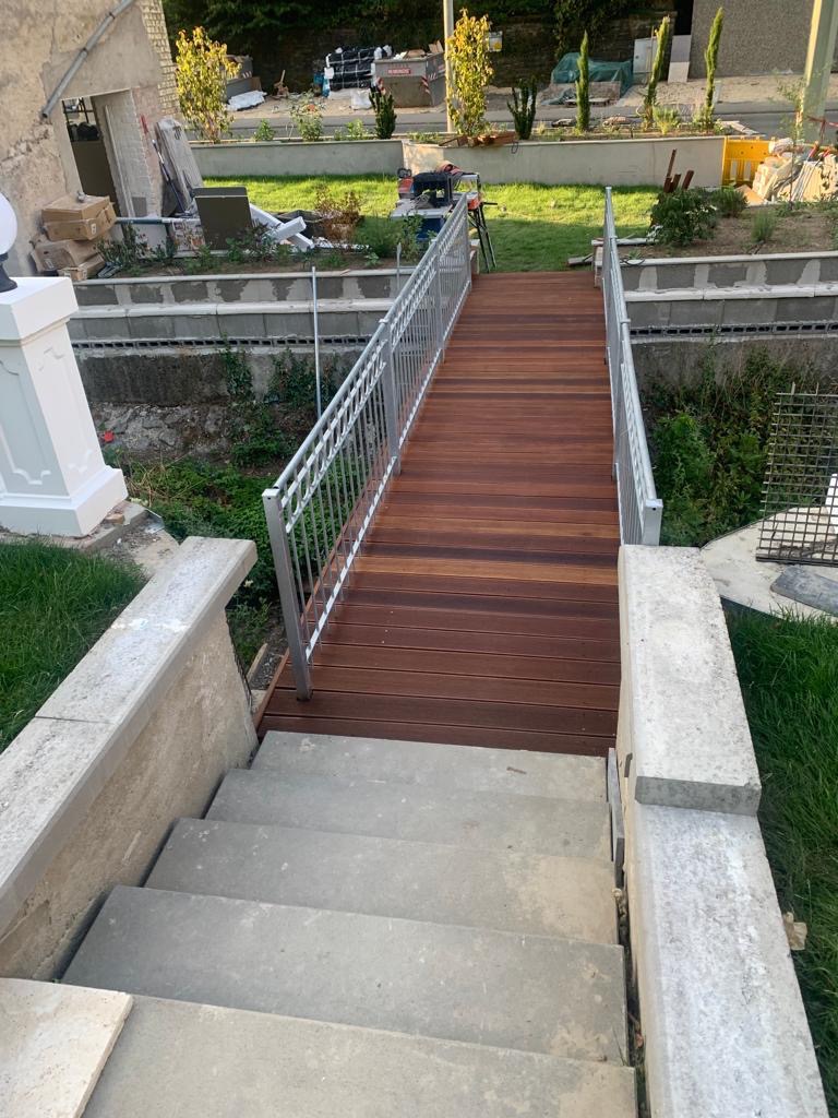 Zewnętrzne schody z drewnianym mostkiem i metalową balustradą, prowadzące do ogrodu z roślinami i elementami dekoracyjnymi. Widok z góry, ciepłe światło.