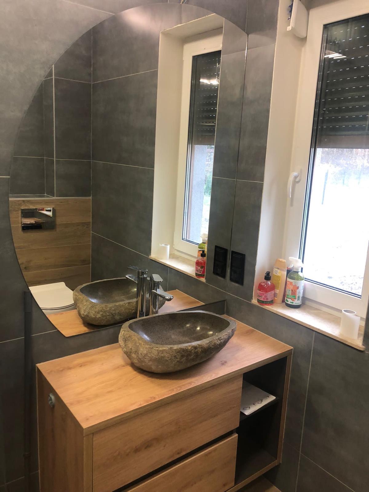 Nowoczesna łazienka z szarą glazurą, drewnianą szafką pod umywalkę z kamienia i owalnym lustrem. Widoczny fragment toalety i okno z roletami.