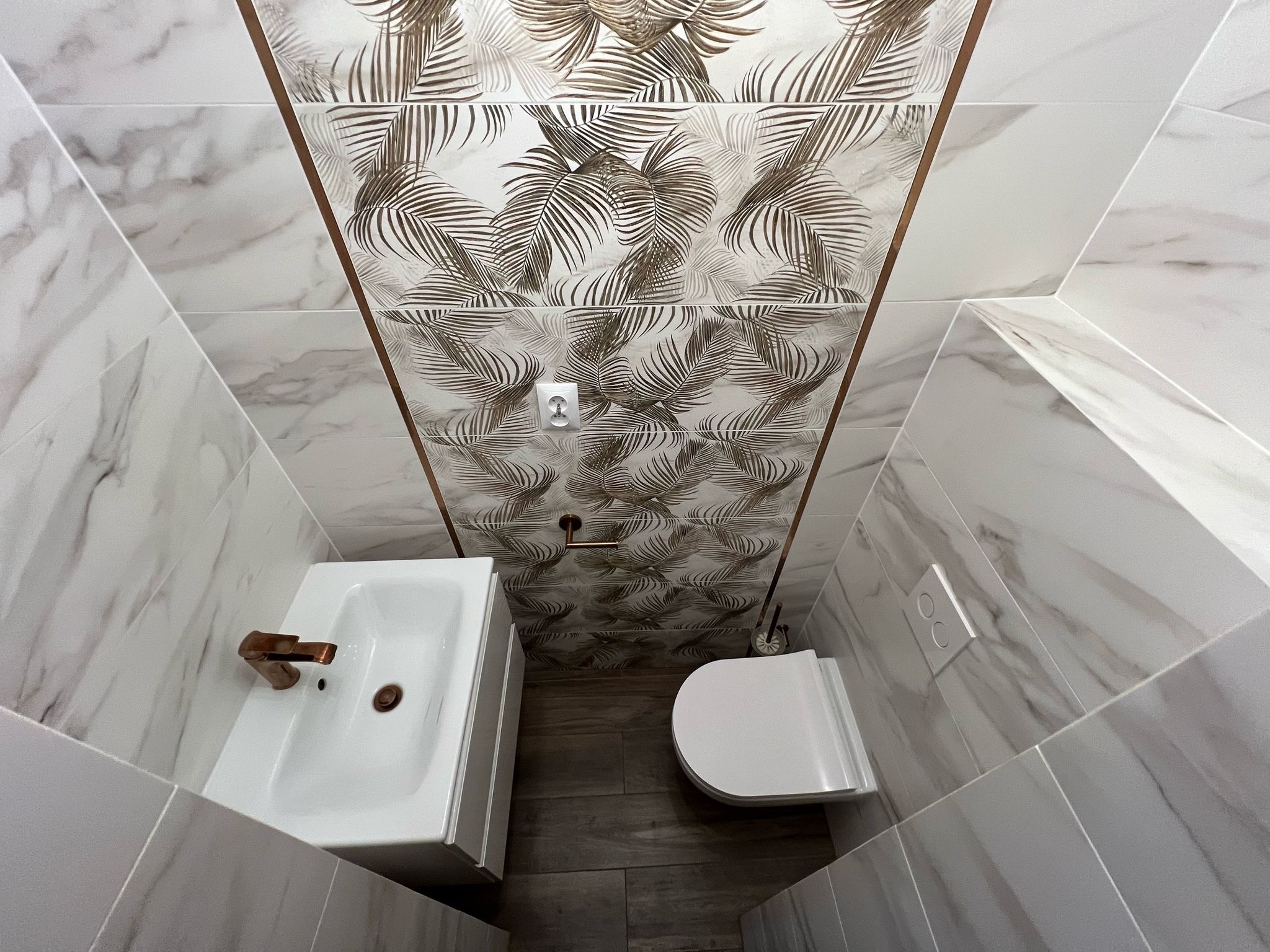 Wykończona łazienka z marmurowymi płytkami, umywalką i toaletą. Na ścianach płytki z motywem liści palmowych, armatura w kolorze miedzi.
