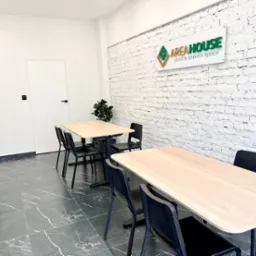 Wnętrze biura agencji nieruchomości z białymi cegłami, stołami, krzesłami, roślinami doniczkowymi i logo firmy AREA HOUSE na ścianie.