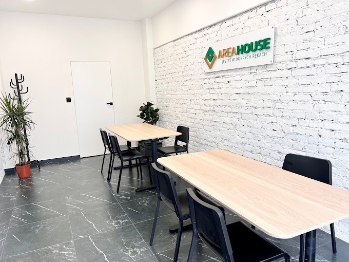 Wnętrze biura agencji nieruchomości z białymi cegłami, stołami, krzesłami, roślinami doniczkowymi i logo firmy AREA HOUSE na ścianie.