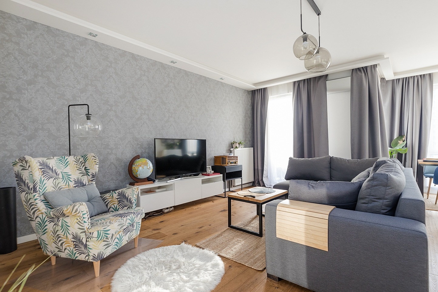 Jasne wnętrze salonu z szarą kanapą, wzorzystym fotelem, drewnianą podłogą i globusem na szafce RTV.