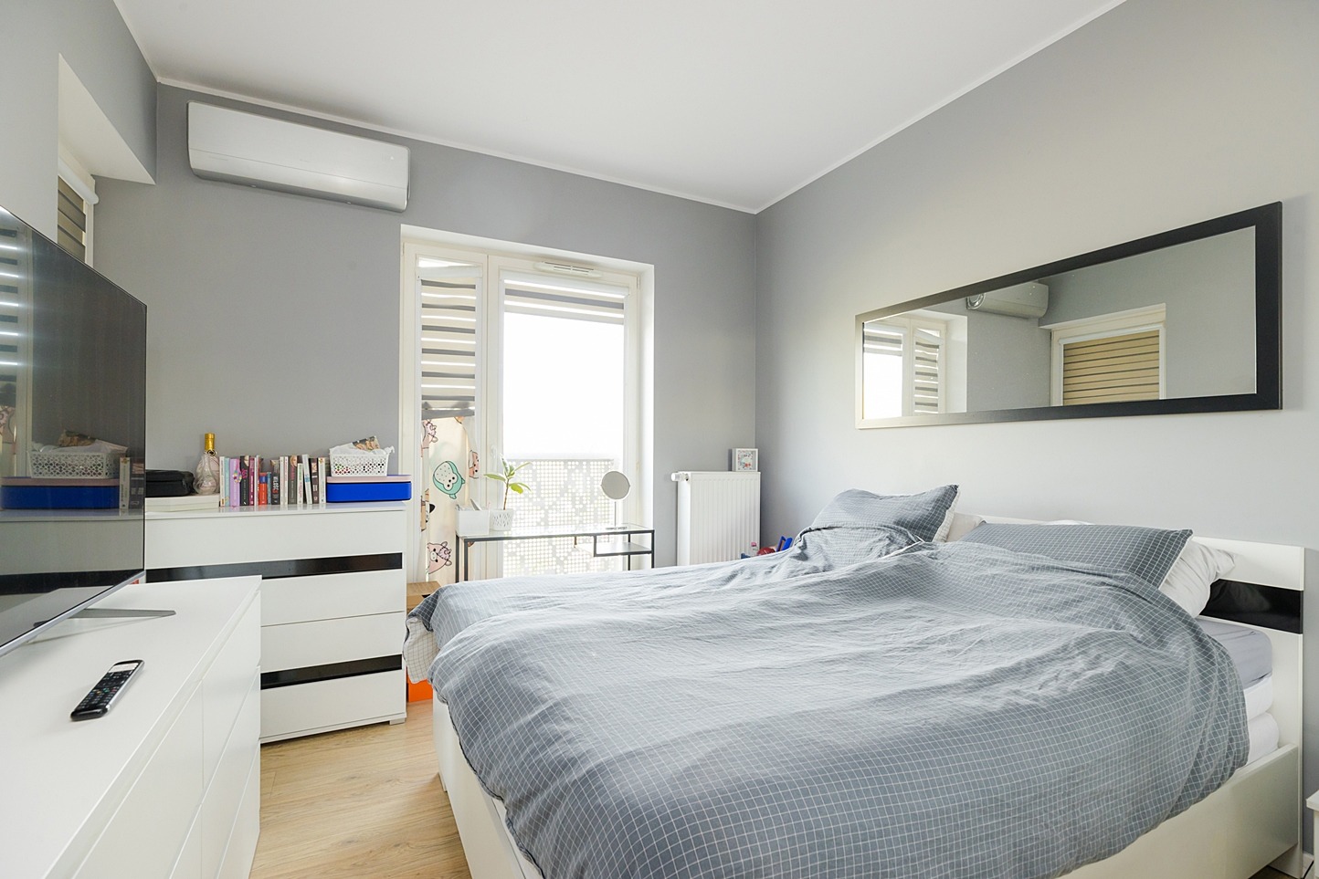 Jasna sypialnia z szarą pościelą w kratę, białymi meblami, dużym lustrem w czarnej ramie i klimatyzatorem na ścianie