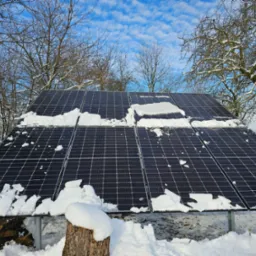 Panele słoneczne częściowo pokryte śniegiem na tle zimowego krajobrazu z drzewami bez liści i błękitnym niebem z chmurami.