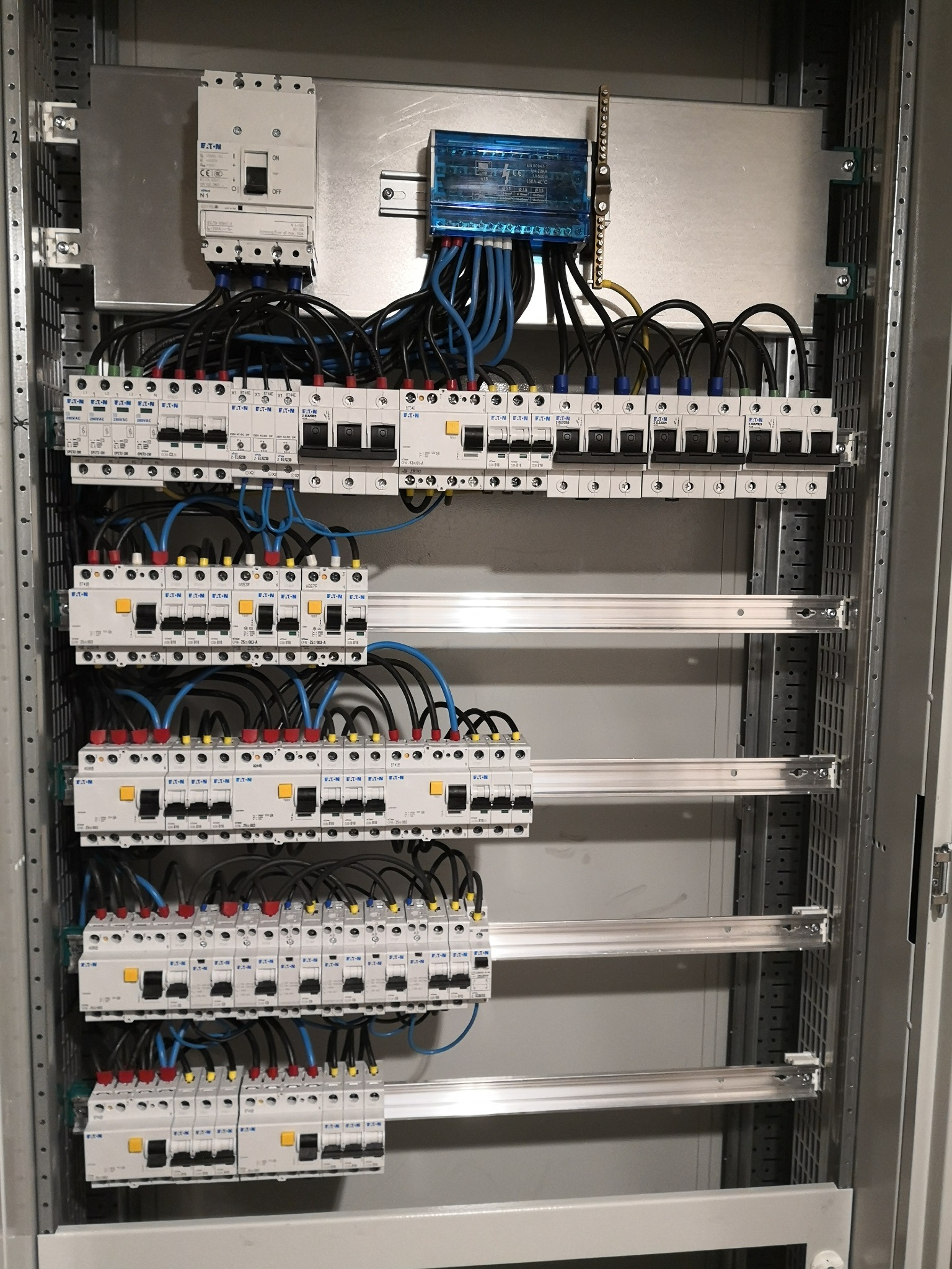 Rozdzielnica elektryczna z zainstalowanymi bezpiecznikami EATON, widoczne przewody w kolorach czarnym i niebieskim, szyny montażowe wewnątrz metalowej obudowy, zabezpieczenie przeciwprzepięciowe...