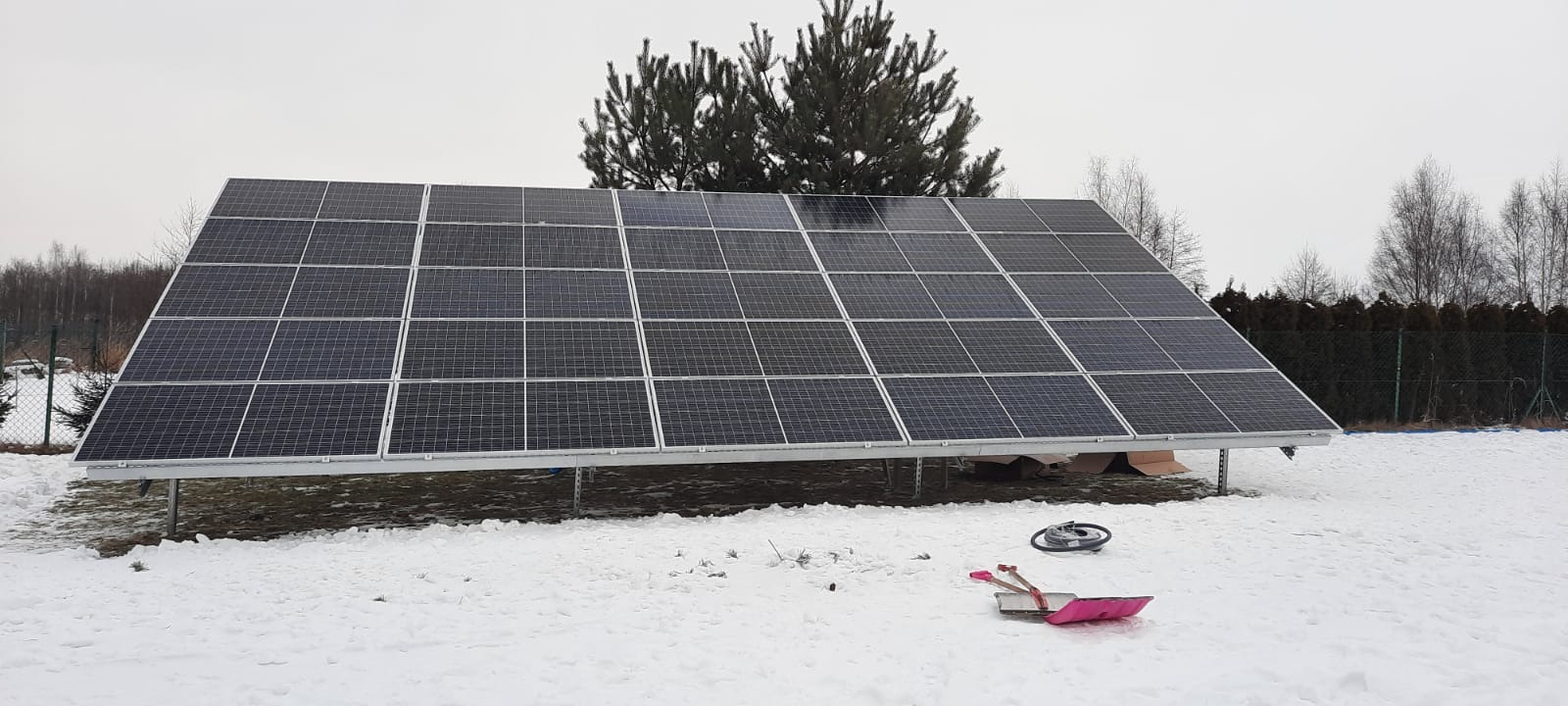 Panel słoneczny na metalowej konstrukcji, ustawiony na zasypanym śniegiem trawniku, z widoczną różową łopatą do śniegu i rozpakowanymi kartonami pod spodem. W tle drzewa i krzewy.