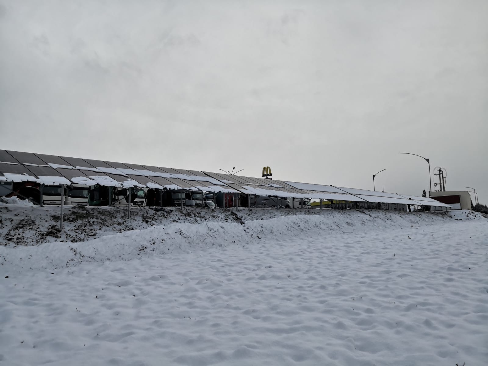 Rząd autobusów zaparkowanych pod zadaszeniem pokrytym panelami słonecznymi, lekko ośnieżonymi, w zimowym krajobrazie z logiem restauracji fast food w tle.