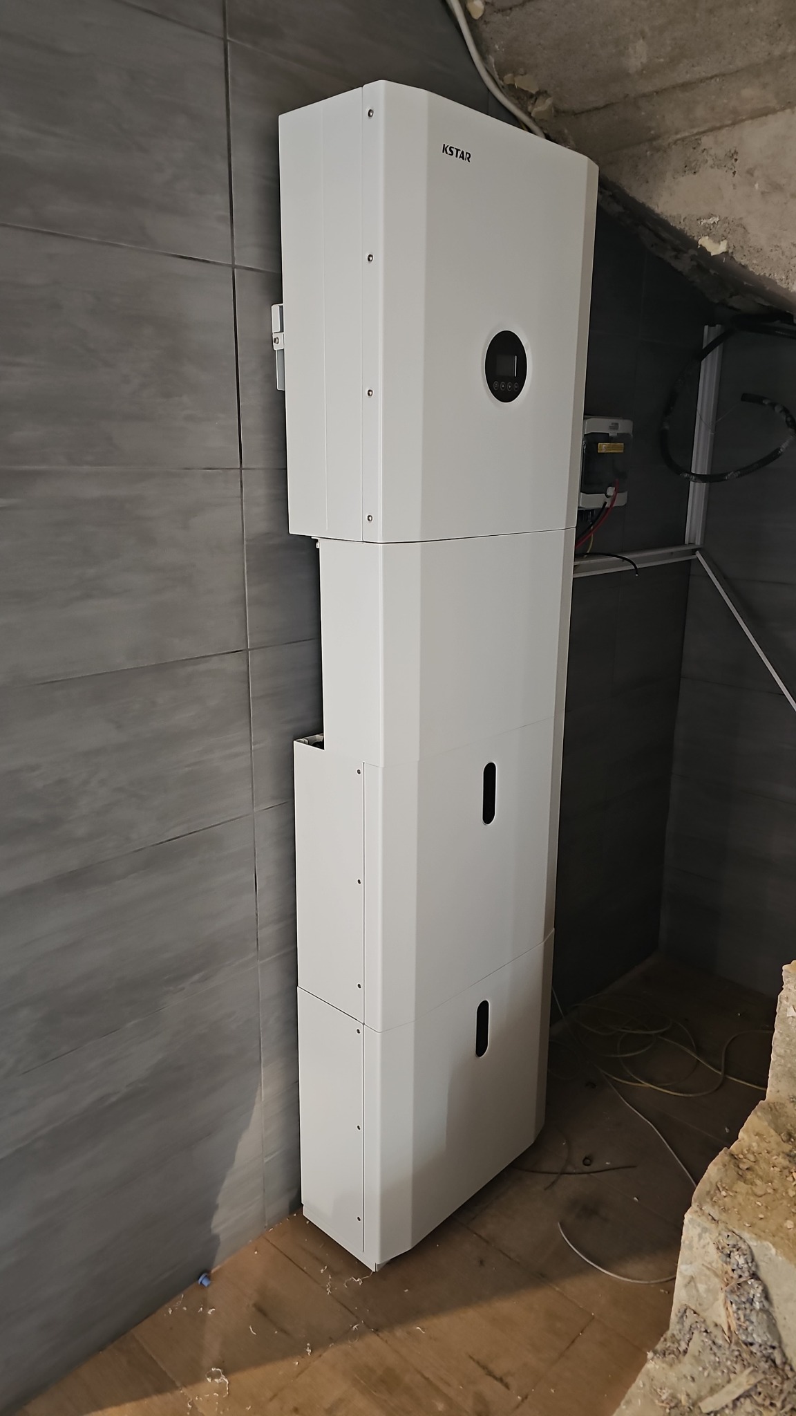 Biała pompa ciepła KSTAR, zamontowana przy szarej, kafelkowanej ścianie, widoczne przewody elektryczne i fragment surowego wykończenia pod sufitem.