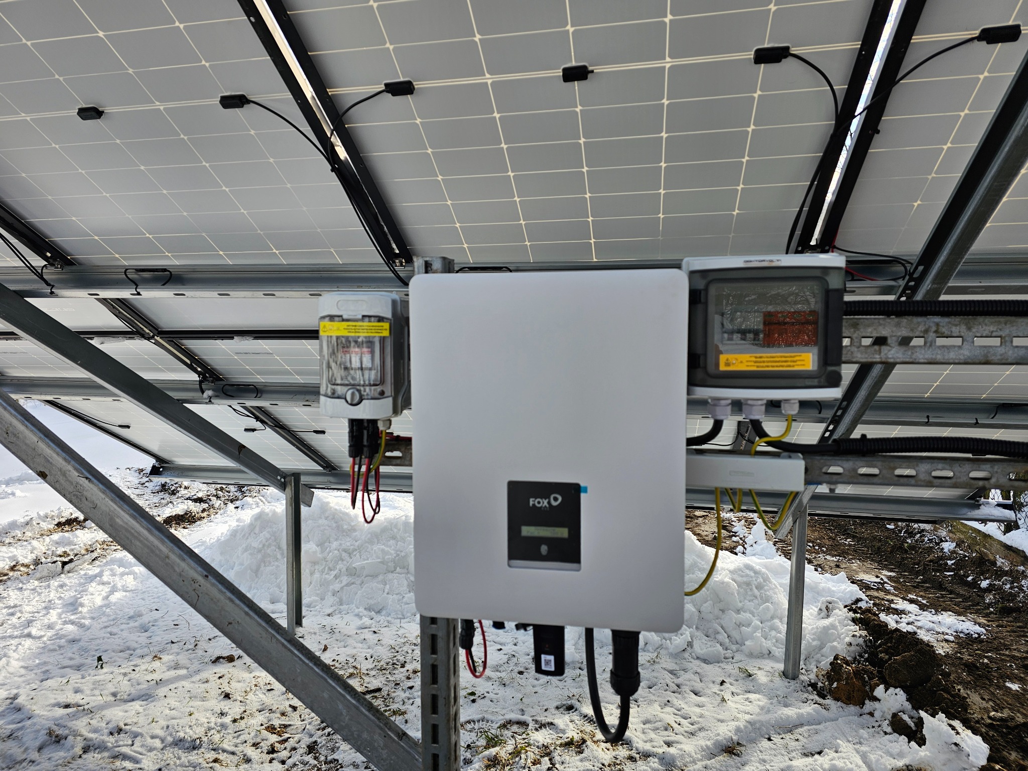 Widok od dołu na panele słoneczne z zamontowanym inwerterem FoxESS i licznikiem energii na tle ośnieżonego terenu.