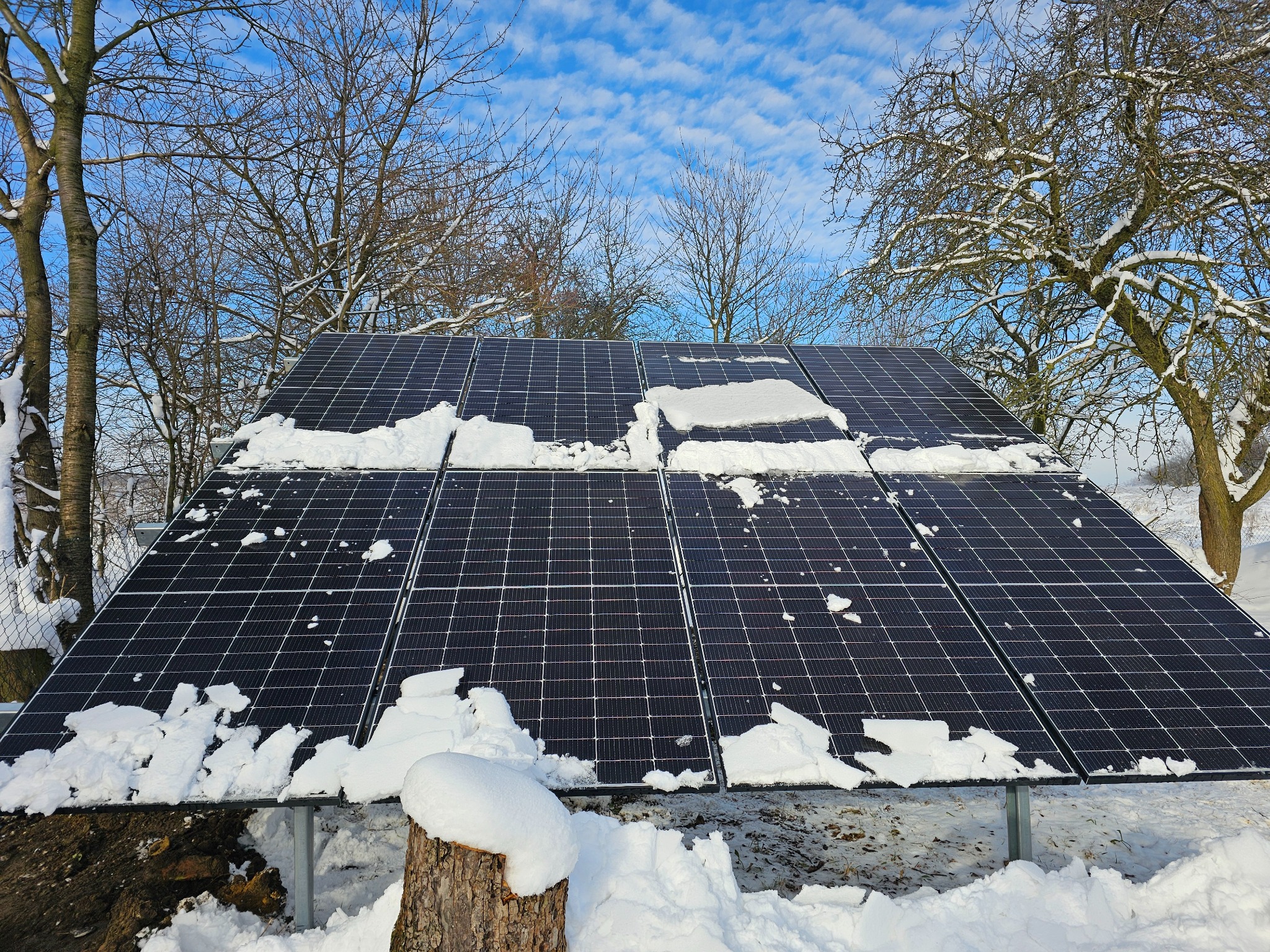 Panele słoneczne częściowo pokryte śniegiem na tle zimowego krajobrazu z drzewami bez liści i błękitnym niebem z chmurami.