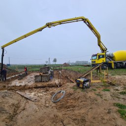 Wytwórnia Materiałów Budowlanych Zdzisława Szczepańska - Żółta betonomieszarka z wysięgnikiem pompuje beton do szalunków fundamentowych, widoczny generator prądu, zbrojenie i pracownicy budowlani w odblaskowych kamizelkach w pochmurny dzień.