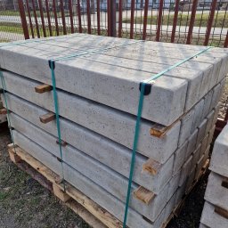 Wytwórnia Materiałów Budowlanych Zdzisława Szczepańska - Paleta z prefabrykowanymi, szarymi elementami betonowymi, spięta zielonymi taśmami, z drewnianymi przekładkami, na tle metalowego ogrodzenia i budynków.