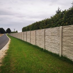 Wytwórnia Materiałów Budowlanych Zdzisława Szczepańska - Długi, szary, betonowy mur o fakturze kamienia, oddzielający jezdnię od trawnika i żywopłotu z tui, w tle pole i drzewa.