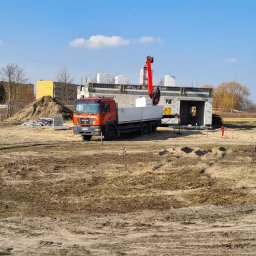 Wytwórnia Materiałów Budowlanych Zdzisława Szczepańska - Pomarańczowa ciężarówka MAN z czerwoną platformą dźwigową dostarcza białe bloczki na plac budowy domu z szarej cegły, widoczny stos szarych kostek brukowych i hałda ziemi na pierwszym planie.