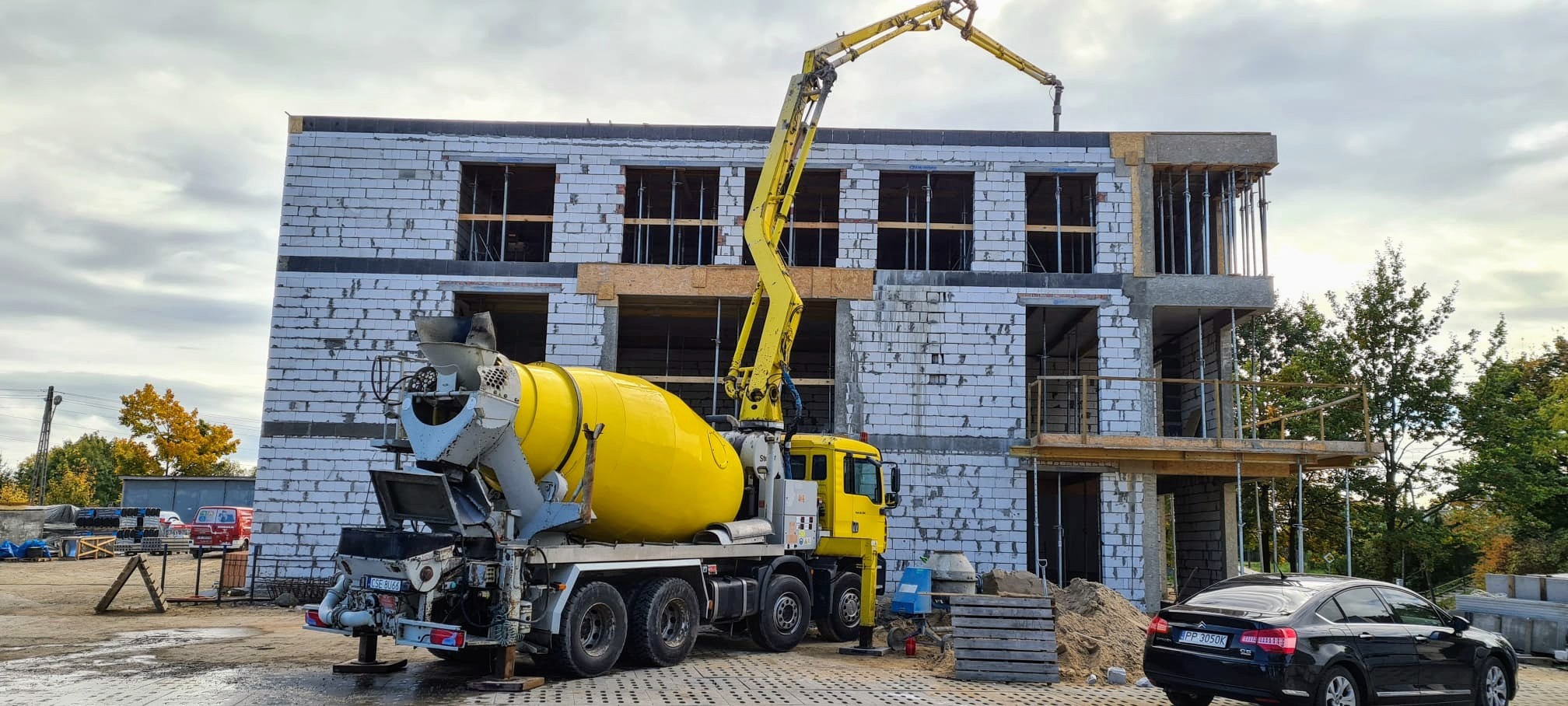 Żółta betonomieszarka z wysięgnikiem podaje beton na budowę dwukondygnacyjnego budynku z białych bloczków, widoczne szalunki i zbrojenia, obok zaparkowany czarny samochód osobowy.
