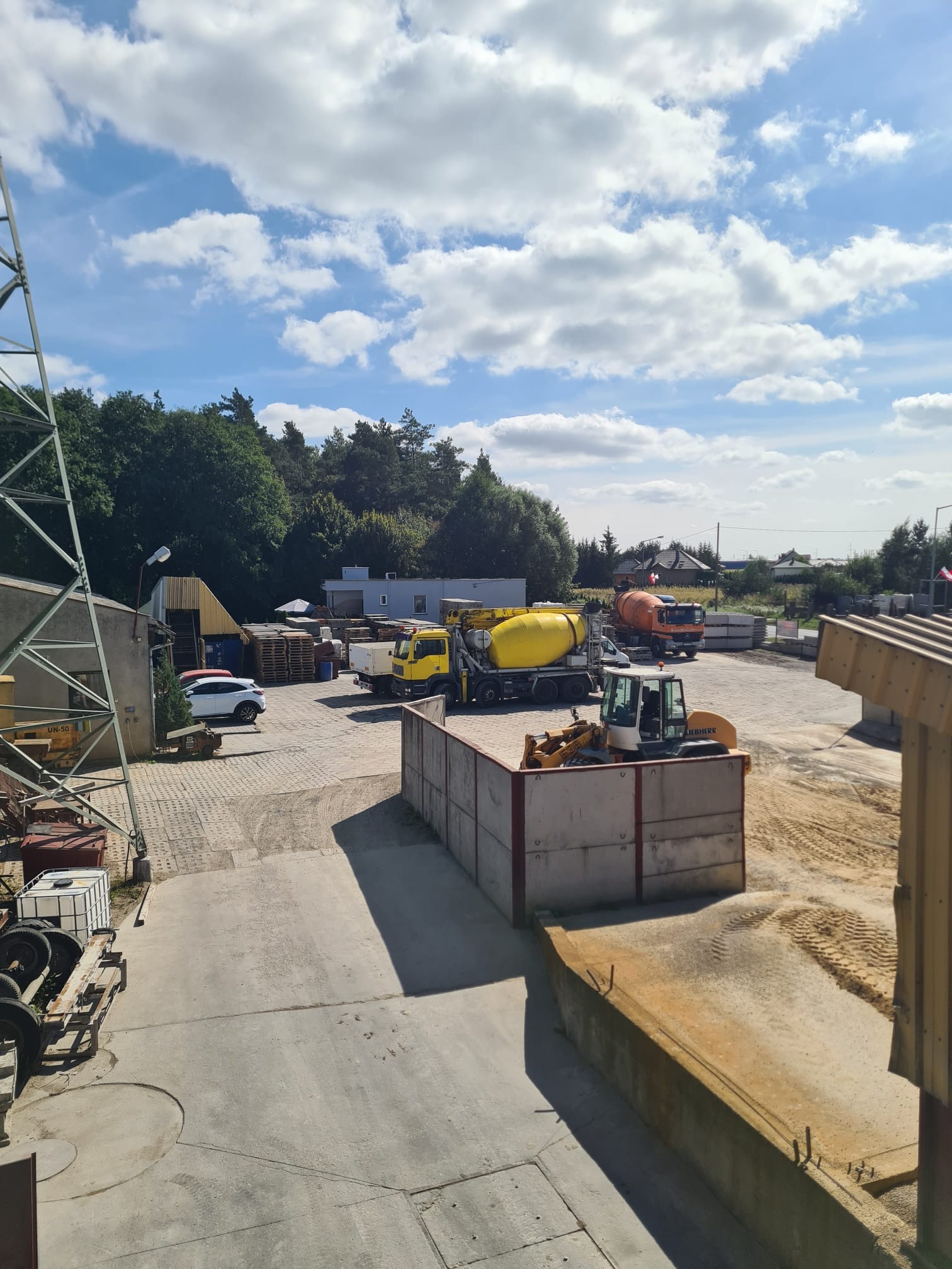 Widok z góry na plac z zapleczem budowlanym, gdzie widać betoniarki, mini ładowarkę, palety i składowane elementy betonowe, a w tle drzewa i błękitne niebo z chmurami.