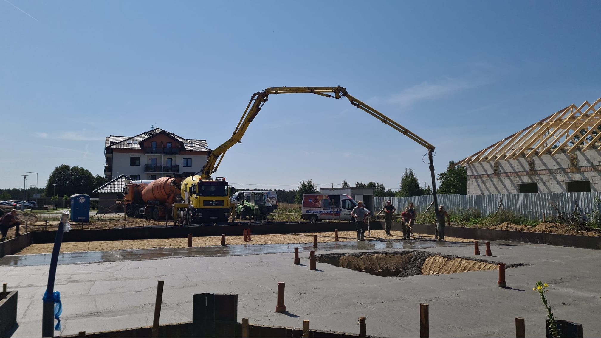 Wylewanie fundamentów domu jednorodzinnego przy użyciu pompy do betonu, widoczny żółty samochód betonomieszarki MAN oraz ekipa budowlana w trakcie pracy.
