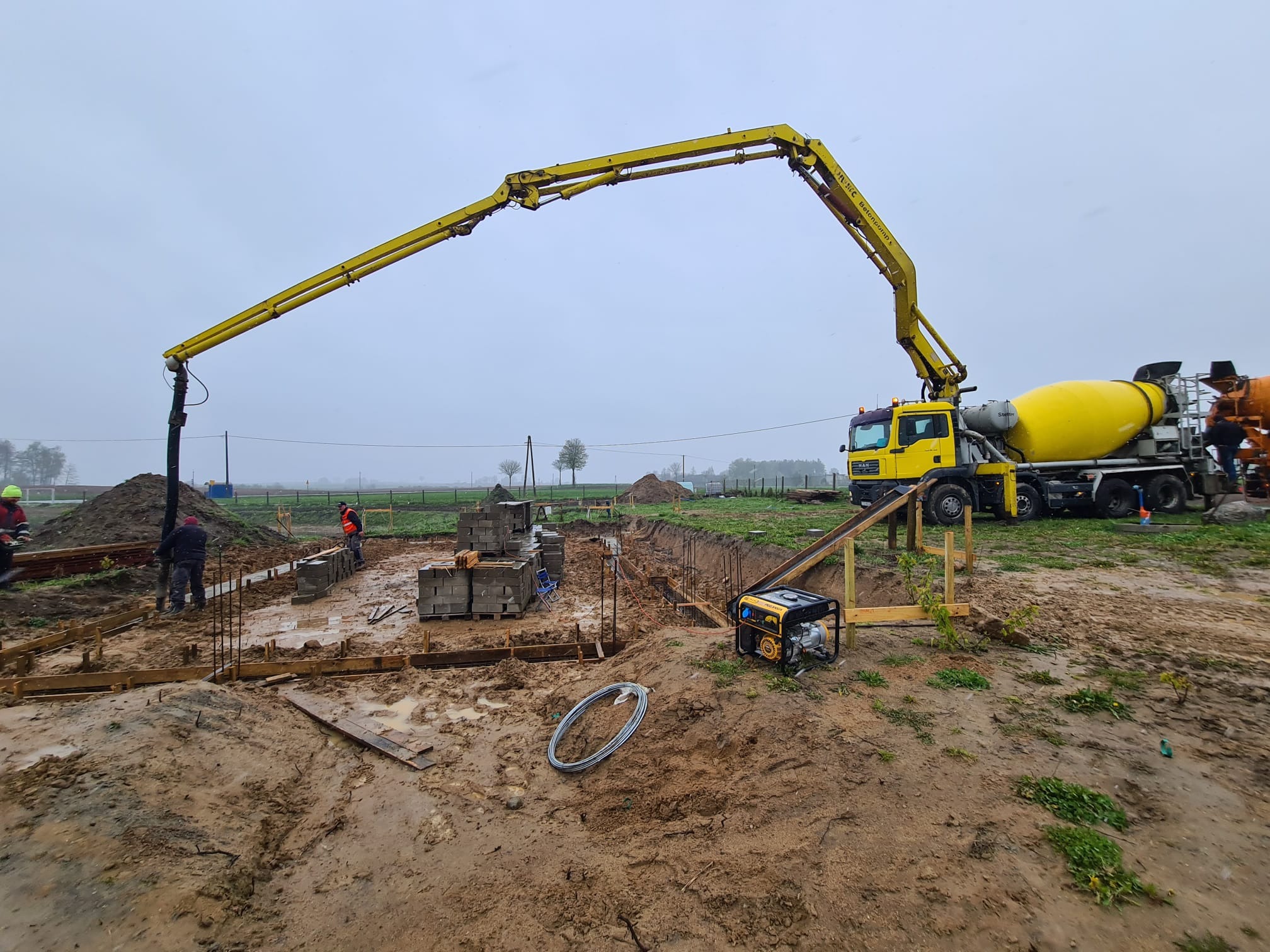 Żółta betonomieszarka z wysięgnikiem pompuje beton do szalunków fundamentowych, widoczny generator prądu, zbrojenie i pracownicy budowlani w odblaskowych kamizelkach w pochmurny dzień.