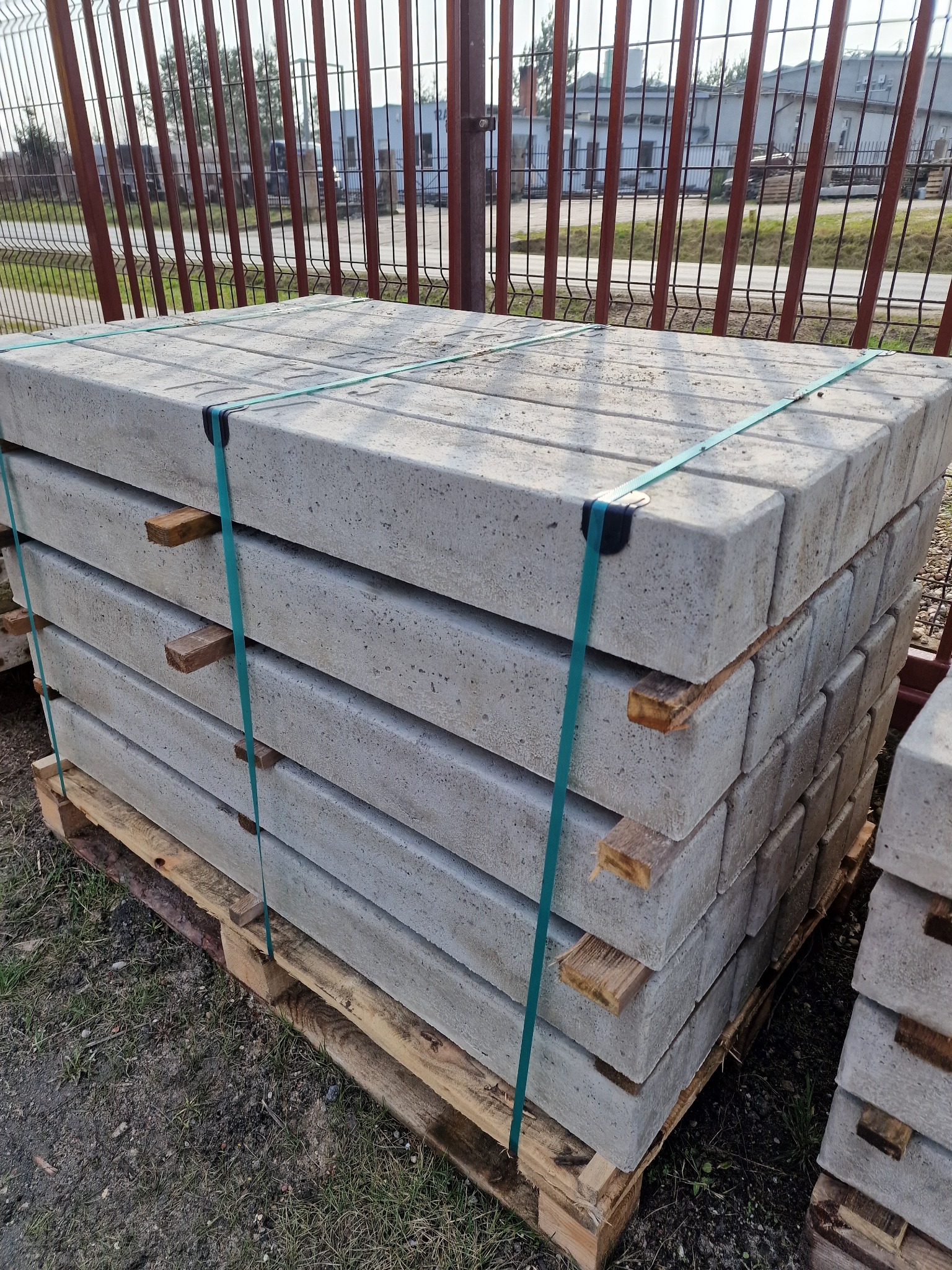 Paleta z prefabrykowanymi, szarymi elementami betonowymi, spięta zielonymi taśmami, z drewnianymi przekładkami, na tle metalowego ogrodzenia i budynków.