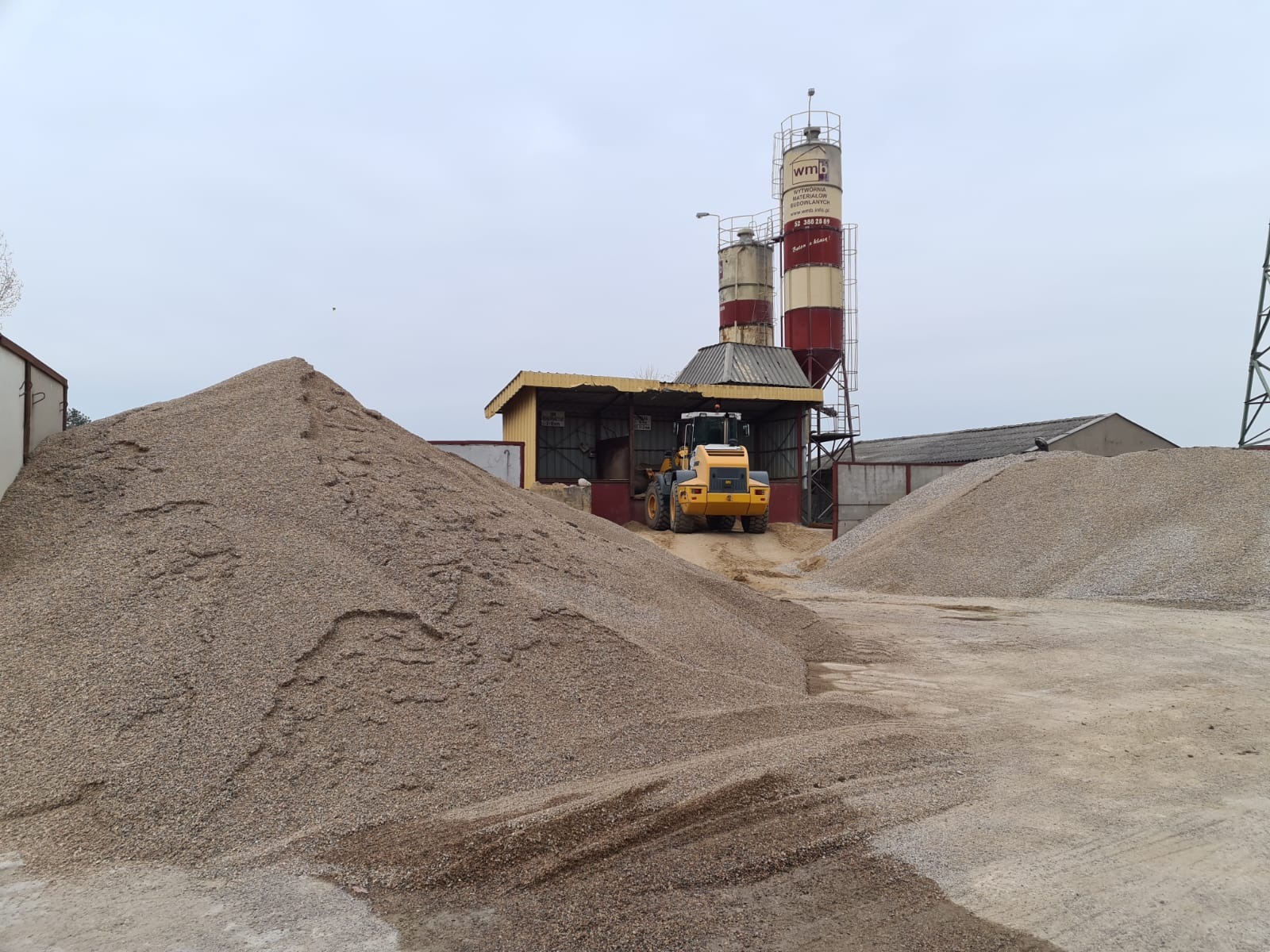 Żwirownia z hałdami kruszywa i żółtą ładowarką na tle silosów pod pochmurnym niebem.