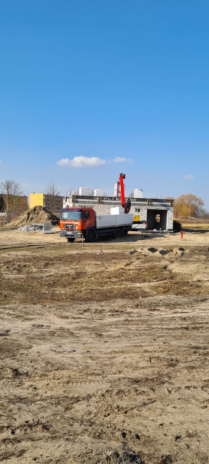 Pomarańczowa ciężarówka MAN z czerwoną platformą dźwigową dostarcza białe bloczki na plac budowy domu z szarej cegły, widoczny stos szarych kostek brukowych i hałda ziemi na pierwszym planie.