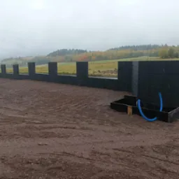 Wznoszenie ogrodzenia z prefabrykat&oacute;w betonowych w pochmurny dzień, widoczny fragment fundamentu i niebieskie rury drenażowe.