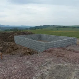 Wzniesiony fundament z bloczków betonowych na terenie budowy, widoczne pryzmy ziemi i oddalony krajobraz pagórkowaty pod pochmurnym niebem.