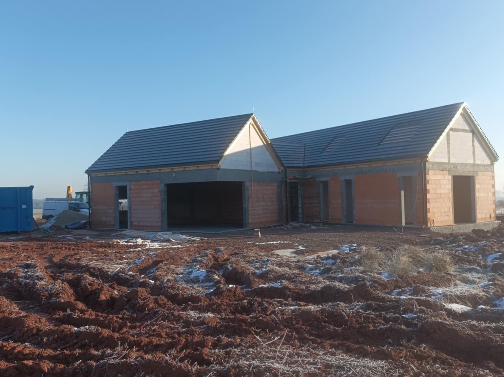 Wznoszony budynek jednorodzinny z garażem w stanie surowym otwartym, widoczne ściany z czerwonej cegły, szara dachówka, teren wokół budynku z nierównościami i resztkami śniegu, w tle błękitne niebo...