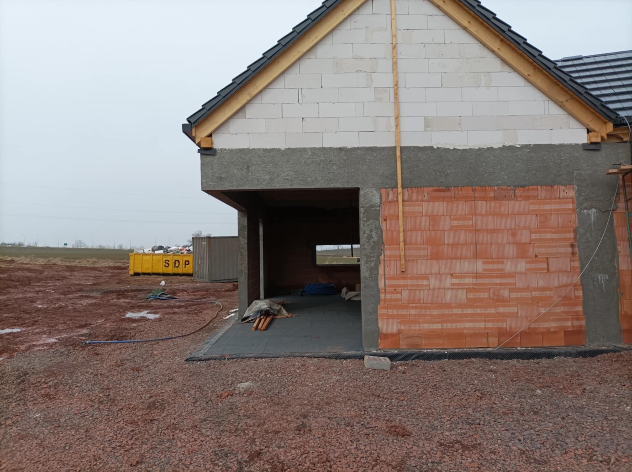 Budowa domu w stanie surowym otwartym: widoczna ściana z czerwonej cegły, białe bloczki na górze, fragment dachu z czarnej dachówki, teren wokół domu z gruzem i błotem, kontener budowlany w tle.