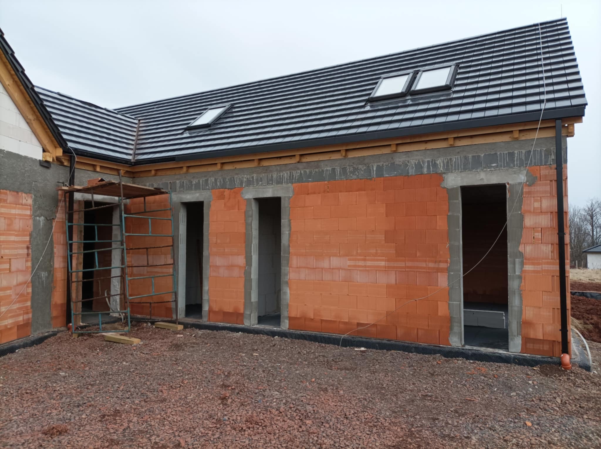 Wznoszone ściany domu z czerwonej cegły ceramicznej z widocznymi otworami okiennymi i drzwiowymi, czarne dachówki na dachu z oknami dachowymi, teren wokół budynku wysypany drobnym żwirem, metalowe...